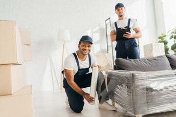 happy indian mover in cap holding stretch film roll near sofa and worker with clipboard