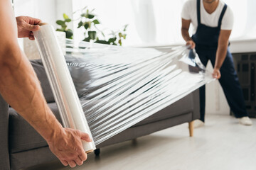 cropped view of movers holding stretch film roll near sofa