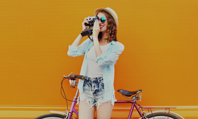 Happy young woman with film camera and bicycle on an orange background