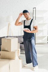 full length of cheerful mover in uniform adjusting cap and holding digital tablet in apartment