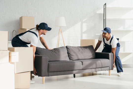 happy multicultural movers in uniform smiling while carrying couch in apartment