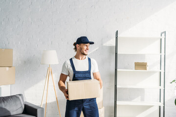 happy mover in cap holding carton box and looking away in apartment