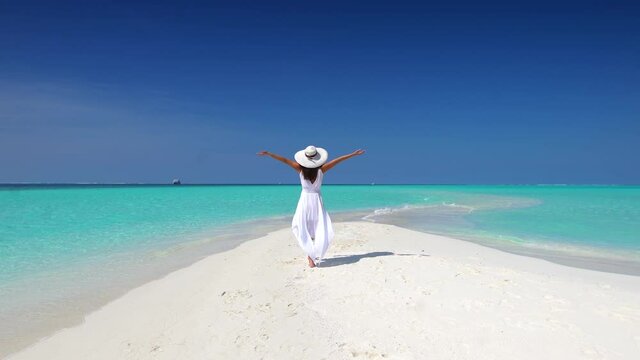 A woman with white hat and outstretched arms walks over a tropical beach towards turquoise ocean and enjoys her exotic getaway