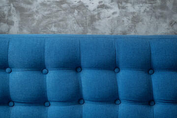 A piece of upholstered furniture with classic Chesterfield pattern with buttons on grey concrete wall background. Modern blue furniture fabric. Elegant luxury sofa upholstery. Interior backdrop