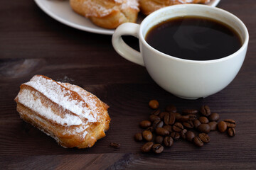 homemade vanilla eclairs or profiteroles on a white plate on a wooden table. copy space. Traditional french tasty dessert on dark background. cup of black coffee with beans.