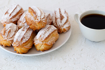homemade vanilla eclairs or profiteroles on a plate on a white table. copy space. Traditional french tasty dessert and cup of black coffee on a background.