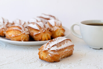 homemade vanilla eclairs or profiteroles on a plate on a white table. copy space. Traditional french tasty dessert and cup of black coffee on a background.