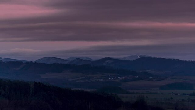 Timelapse of the peaks of the Pustevny mountains covered with snow in the background is a sunset and fast moving clouds across the sky.
