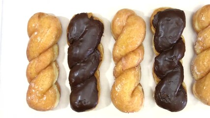 4K HD video panning across row of chocolate covered and plain glazed twist donuts on parchment paper over light wood table.
