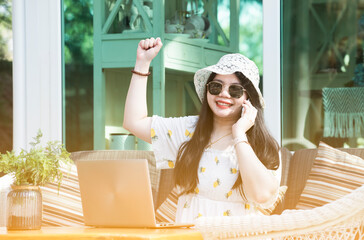 Young woman in sunglasses talking by phone and laptop siting on sofa at terrace in front of house. Work form home. Woman working business online with smart phone and laptop in comfortable mood at home