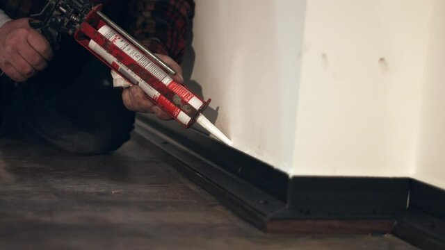 Grout sealent out of silicon with a white color being applied on a skirting base board in a new apartment by a crafts person with a cartridge gun on a black wooden floor. Shot in 4K.