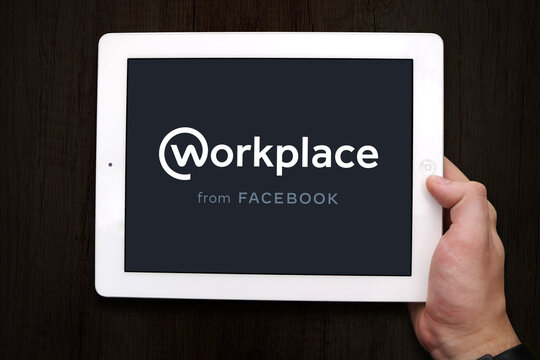 A Hand Holding An IPad Or Tablet Device With Workplace (by Facebook) Logo Displayed In Full Screen. Workplace Is An Enterprise Connectivity Platform That Use Groups, Instant Messaging And News Feed.