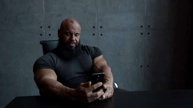 a muscular bald man sits at home at a Desk and uses a smartphone, checks messages and smiles.