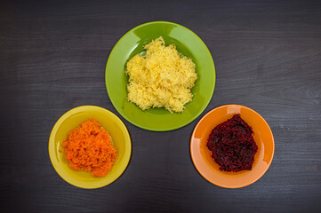 A set of ingredients for making salads. Mashed boiled carrots, beets, potatoes in plates on a dark wooden table. Flat lay