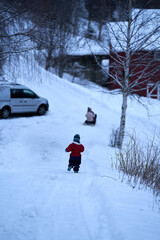 A little boy running after his sisters in the winter. The girls are riding a sleigh or toboggan. 