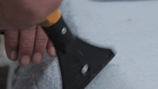 Person hand cleans ice from white electric vehicle hood with snow brush of yellow and black colours. Concept seasonal disaster