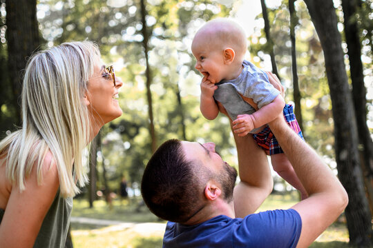 Photography of family with one child in nature. Dad is holding baby in air, while mom is standing behind him and coo to baby. Boy is smiling back. Weekend family time.