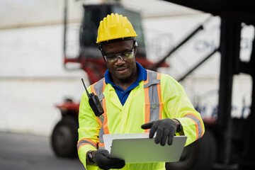 African technician dock worker in protective safety jumpsuit uniform and with hardhat and use digital tablet at cargo container shipping warehouse. transportation import,export logistic industrial