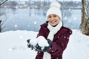 A happy woman wearing a white knitted woolen hat and scarf smiles and holds snowballs on the hands. Portrait on a beautiful lake background. Snowy Day.Falling snow