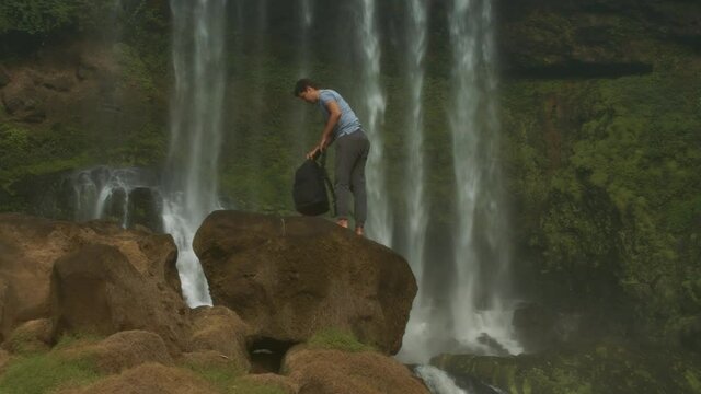 Guy tourist stands on huge brown rock takes off backpack drinks water from bottle against fantastic waterfall