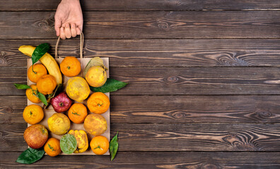 Various ripe, raw fruits in package. Woman's hand holds package, dark restored wooden background. Top view. Food paper bag idea of zero waste. Food delivery, purchase or donation concept. Copy space