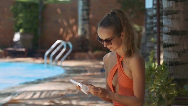 Closeup slim blond girl in orange bikini smears arms with sunscreen sitting in shade on sunny day