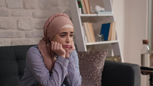 Woman in a turban sitting on the sofa at home, with her head on her hands.Portrait of thoughtful woman in headscarf sitting on sofa at home, resting her head on her hands and looking away with sad fac