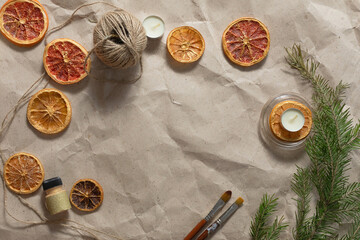 A flat lay of dry slices of orange, lemon, grapefruit, green pine branch, cone, brush, tablet candles are on a kraft beige paper table. Handmade Christmas decoration and candle making concept.