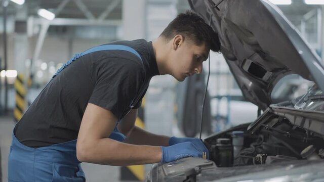 Side view of young brunette Caucasian auto mechanic looking at car engine in open hood and thinking. Portrait of concentrated service man examining vehicle breakage in repair shop.