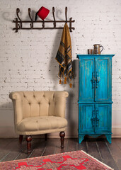 Fototapeta na wymiar Classic beige armchair with wooden legs, vintage turquoise cupboard, wall hanger, and ornate scarf on white bricks wall and grunge wooden parquet with red decorated carpet