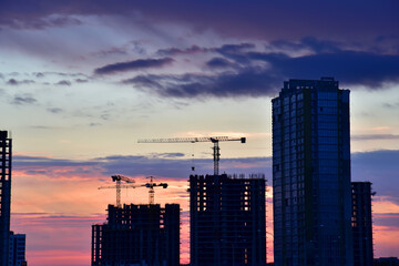 Silhouettes of tower cranes on awesome sunset background. Preparing to pour a bucket of concrete into formwork. Construction a multi-storey residential building. Small sharpness, possible granularity