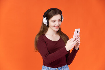 Portrait of smiling young woman in big headphones using mobile phone over orange background