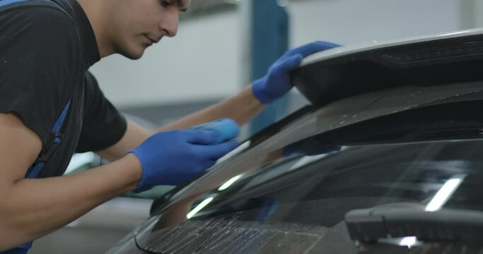 Close-up of handsome young service man cleaning windshield of vehicle. Portrait of professional Caucasian repair shop employee rubbing glass indoors. Cinema 4k ProRes HQ.