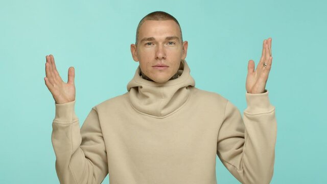 Slow motion of caucasian man with tattoos and piercing, raising hands up and asking why, standing bothered and looking at camera, standing over turquoise background