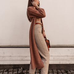 Fashionable young woman in stylish beige trousers in long vintage coat with leather brown trendy...