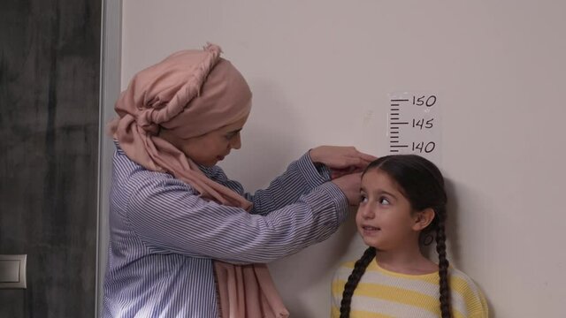 Mother in a turban measuring her daughter's height near a white wall.Young woman is measuring the height of her daughter.