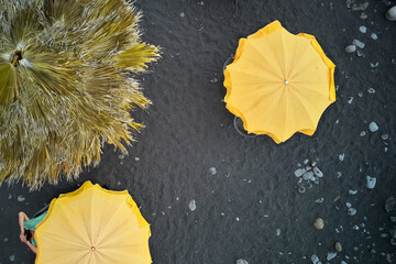 Yellow umbrellas against the background of black volcanic sand. A man is lying under one of the umbrellas. Shooting from a drone.