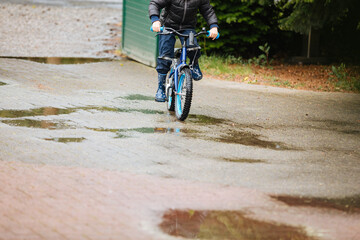 Boy with rain jacket and rubber boots rides a bicycle through puddles. Children play on rainy days
