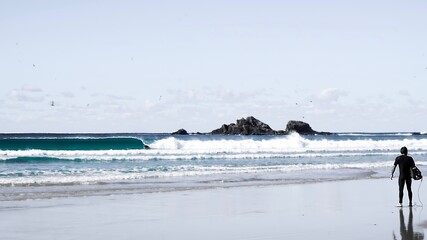 A male surfer holding a surfboard looks on as a wave breaks perfectly infront of him - Byron Bay