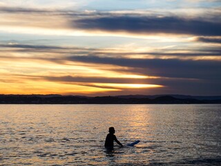 A silhouette of a male surfer sat on his board as the sun sets behind him - Gold Coast, Australia