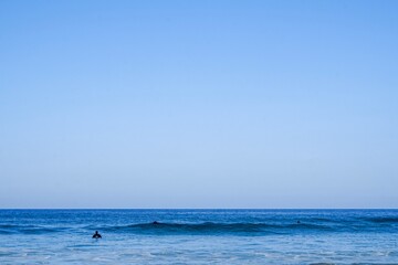 Obraz premium A minimalistic surf line up. Blue skies and blue waters with a small group of surfers sat on thier boards, Cornwall, UK