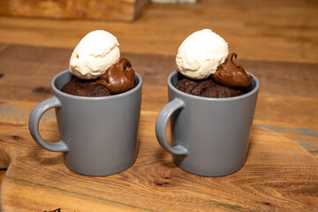 A cup of chocolate cake that has been made in a microwave with chocolate source and ice cream on a wooden kitchen work top