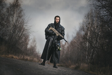 Post apocalypse soldier in the black raincoat and with rifle is standing on the abandoned road.