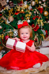 Beautiful little girl in a red dress sits under a Christmas tree and holds a gift box