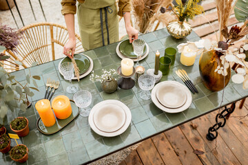 Young female putting herbs on a plates, decorating dinning table in natural Boho style in green tones outdoors