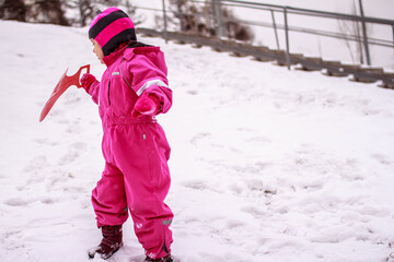 Fototapeta na wymiar Close up of little Caucasian girl wearing pink winter overall holding a snow saucer in winter outdoors . Winter outdoor activities concept. Vertical