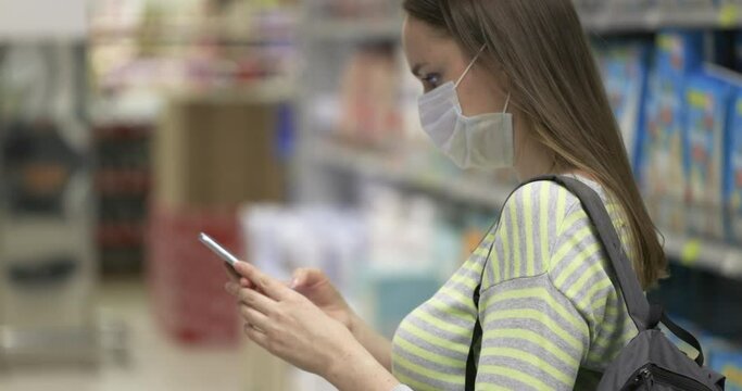 The buyer takes pictures on a smartphone in the store. A woman in a yellow and gray sweater and a protective mask makes purchases in the supermarket. Daily lifestyle, slow motion. 