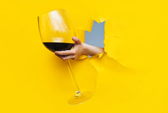 A left woman's hand emerges through a torn hole in yellow paper with a large glass of red wine. The concept of alcoholism, drunkenness and hangover. Copy space.