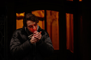 Handsome stylish young hipster man smoking cigarette on the street in winter, looking away. Portrait of beautiful guy dressed casually. Vintage European city, lights on background. Soft focus.