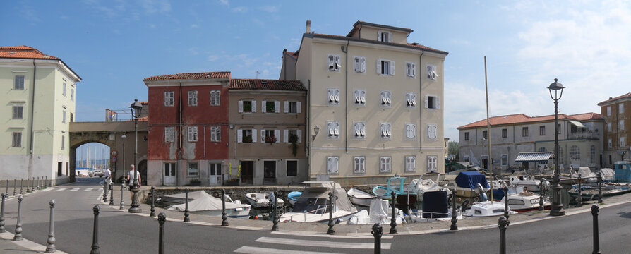Mandracchio in Muggia : panorama of the small harbor inside the village and surrounded by ancient walls and colorful buildings. There are colored boats moored and reflected in the water.